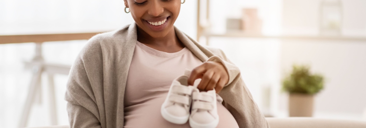 A smiling pregnant lady holding baby shoes against her tummy for postpartum bleeding embolisation