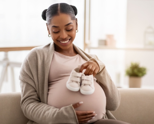 A smiling pregnant lady holding baby shoes against her tummy for postpartum bleeding embolisation